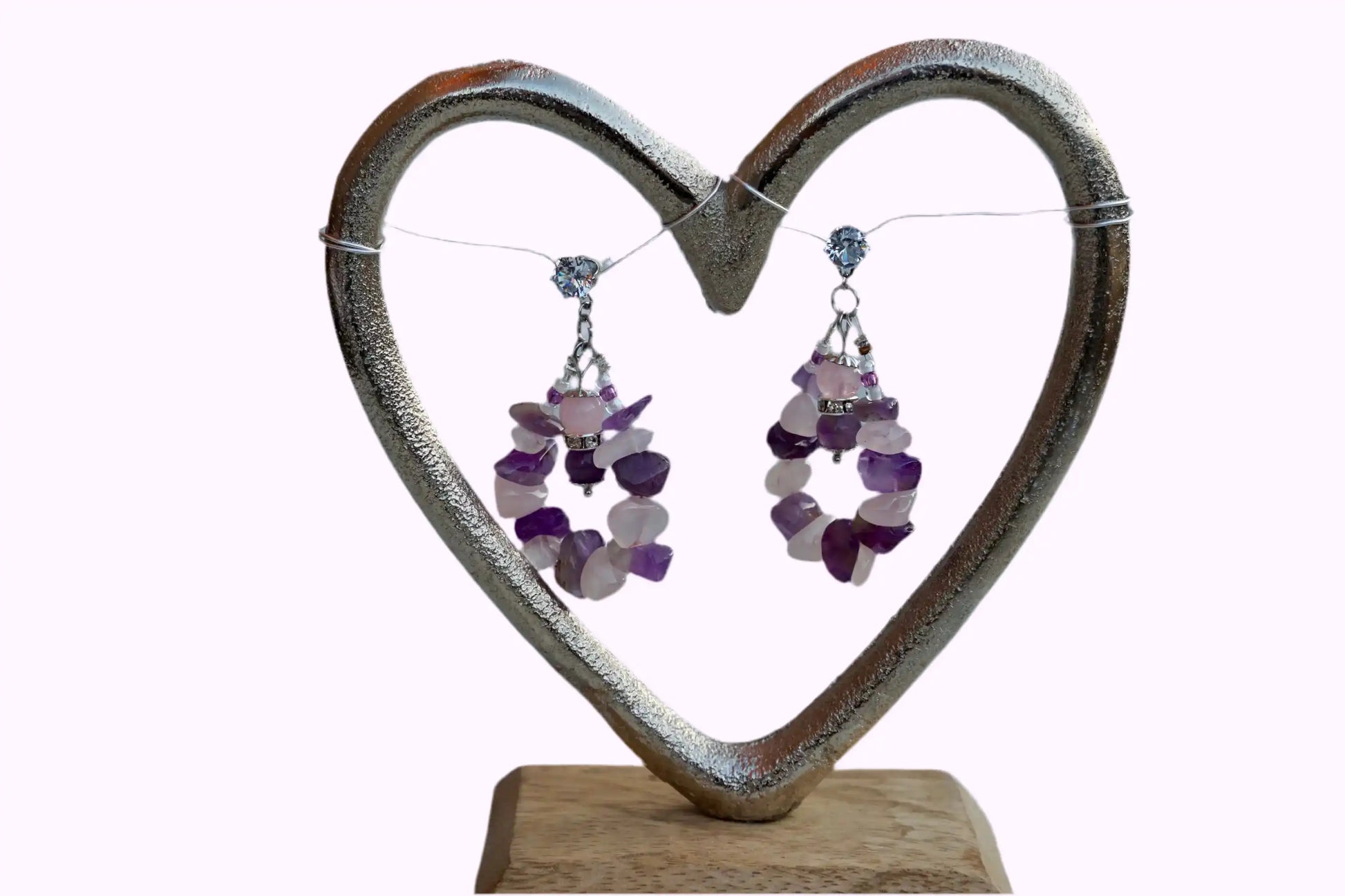 Herzförmiger Schmuckhalter aus Metall mit zwei Ohrringen aus Amethyst und Rosenquarz Steinen, auf Holzsockel.