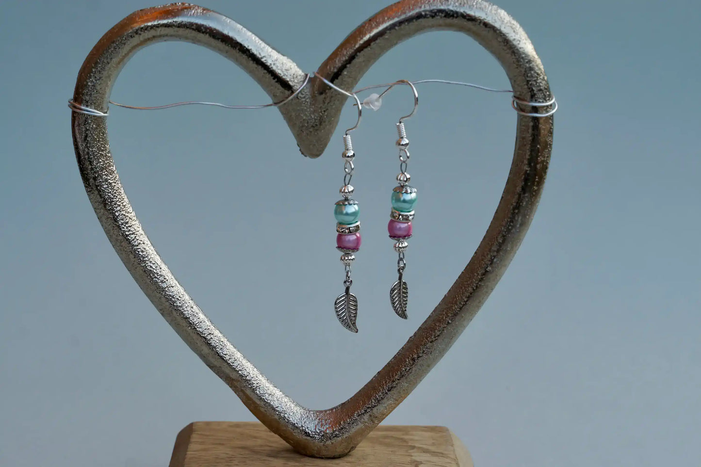 Ohrringe mit silberfarbenen Haken, zwei Perlen in Türkis und Rosa sowie einem silberfarbenen Blattanhänger aufgehängt an einem herzförmigen Metallständer.