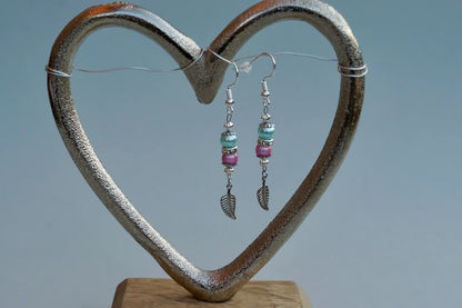 Ohrringe mit silberfarbenen Haken, zwei Perlen in Türkis und Rosa sowie einem silberfarbenen Blattanhänger aufgehängt an einem herzförmigen Metallständer.