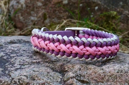 Geflochtenes Hundehalsband aus lila, rosa und grauen Bändern mit silberner Metallschnalle und Löchern zur Größeneinstellung auf einem Stein