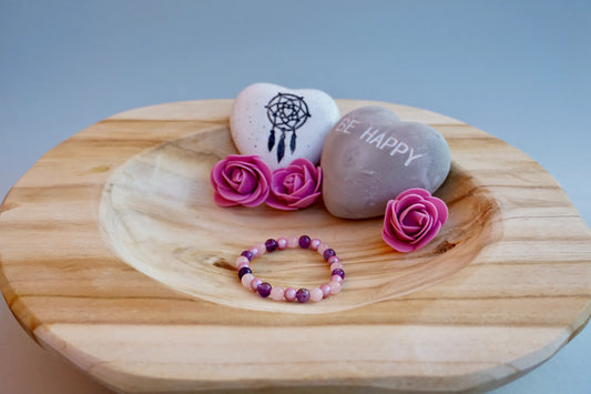Rundes Armband aus Rosenquarz, Amethyst und rosa Acrylperlen auf Holzschale, daneben zwei herzförmige Steine mit Schrift und Traumfänger sowie drei rosa Rosen aus Stoff.