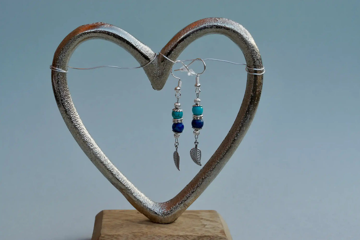Ohrringe mit silberfarbenen Haken, zwei Edelsteinperlen in Turquenit und Sodalith sowie einem silberfarbenen Blattanhänger aufgehängt an einem herzförmigen Metallständer.