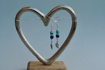Ohrringe mit silberfarbenen Haken, zwei Edelsteinperlen in Turquenit und Sodalith sowie einem silberfarbenen Blattanhänger aufgehängt an einem herzförmigen Metallständer.