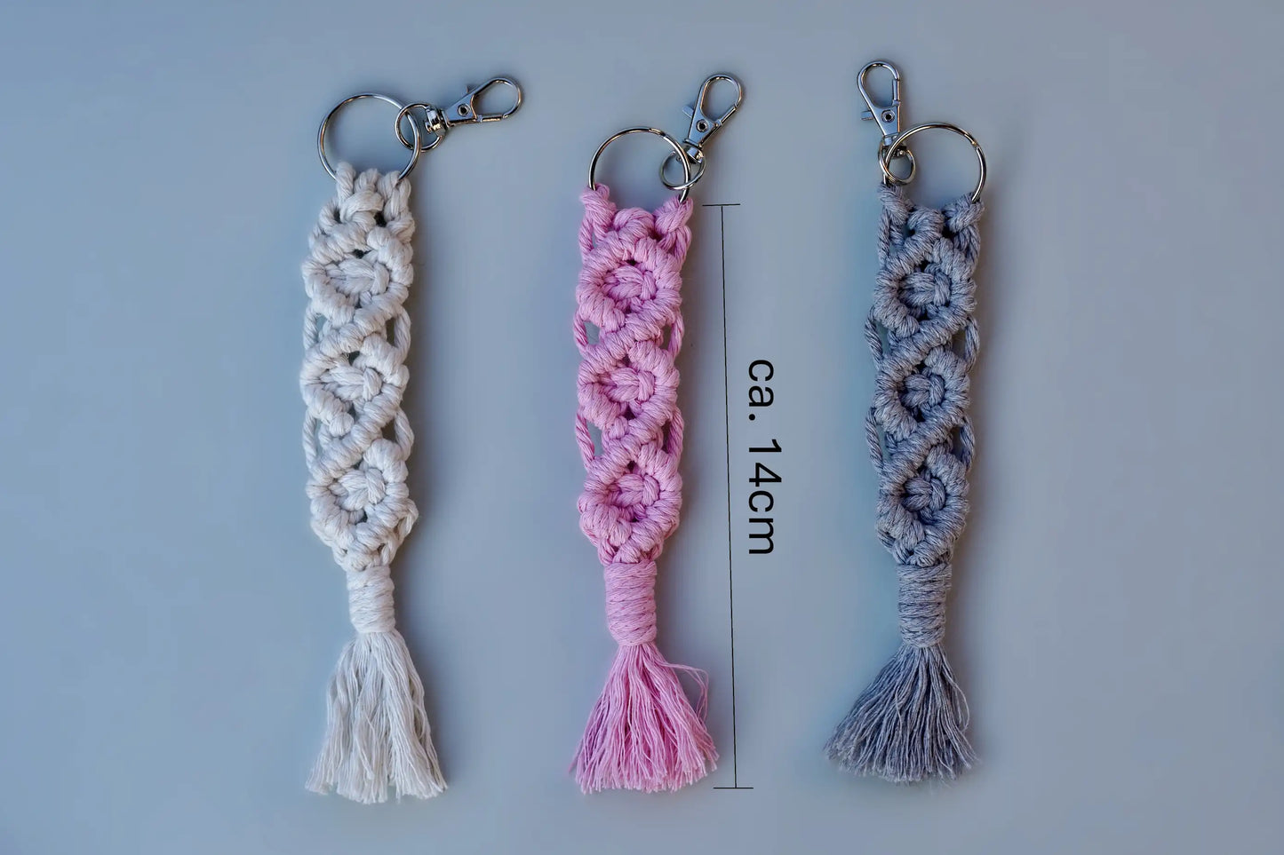 Drei Makramee-Schlüsselanhänger in Weiß, Rosa und Grau mit Knotenmustern und Fransen, jeweils mit Metallring und Karabinerhaken mit Maßstab
