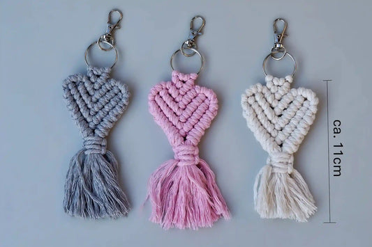 Drei Makramee-Schlüsselanhänger in Grau, Rosa und weiß mit Herzknotenmuster und Fransen, jeweils mit Metallring und Karabinerhaken mit Maßstab