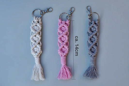 Drei Makramee-Schlüsselanhänger in Weiß, Rosa und Grau mit Knotenmustern und Fransen, jeweils mit Metallring und Karabinerhaken