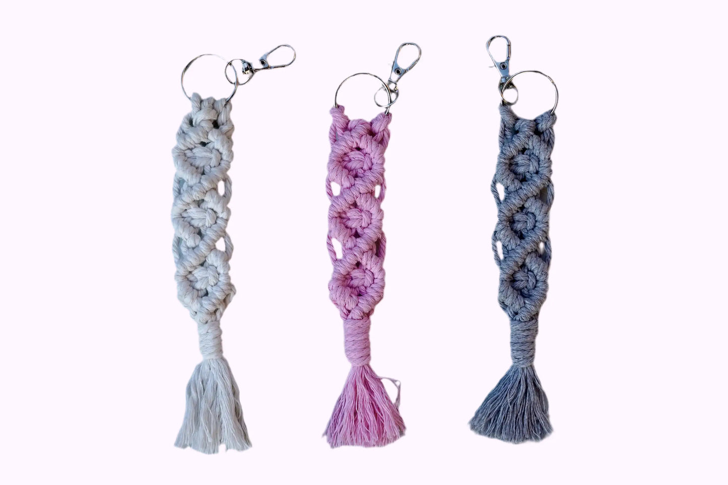 Drei Makramee-Schlüsselanhänger in Weiß, Rosa und Grau mit Knotenmustern und Fransen, jeweils mit Metallring und Karabinerhaken