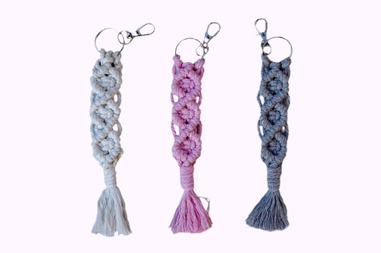 Drei Makramee-Schlüsselanhänger in Weiß, Rosa und Grau mit Knotenmustern und Fransen, jeweils mit Metallring und Karabinerhaken