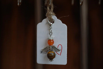 Anhänger in Form eines Schutzengels mit Karneol- und Tigeraugenperlen, silberfarbene Flügel, an einem Band befestigt.