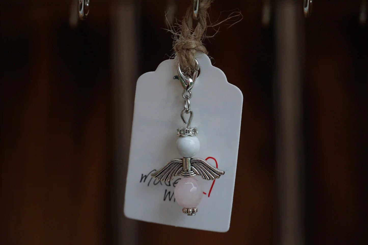 Anhänger in Form eines Schutzengels mit Magnesit- und Rosenquarzperlen, silberfarbene Flügel, an einem Band befestigt.