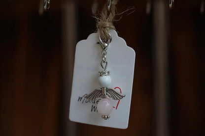 Anhänger in Form eines Schutzengels mit Magnesit- und Rosenquarzperlen, silberfarbene Flügel, an einem Band befestigt.