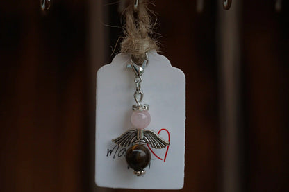 Anhänger in Form eines Schutzengels mit Rosenquarz- und Tigeraugeperlen, silberfarbene Flügel, an einem Band befestigt.