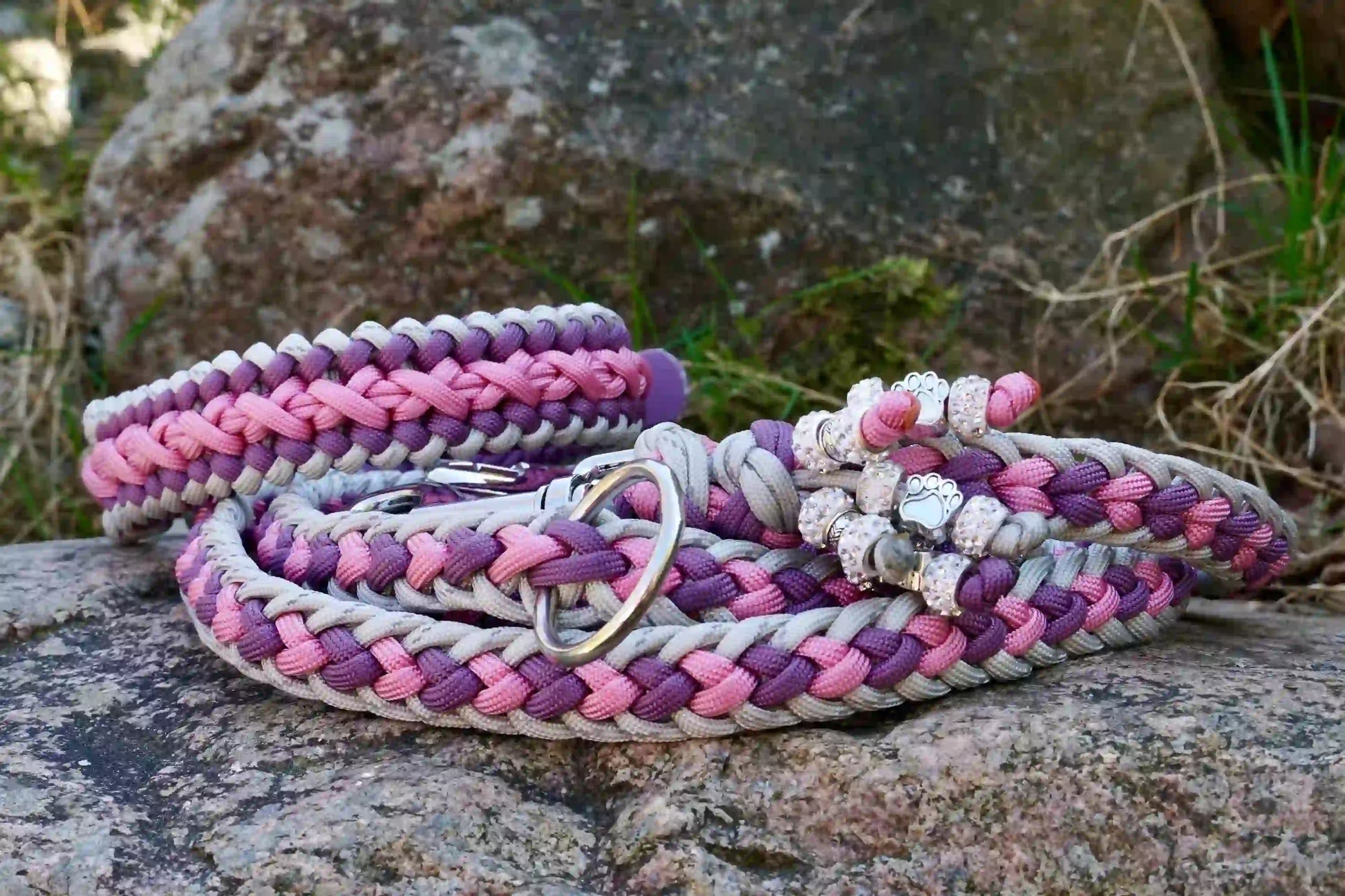 Geflochtene Hundeleine und Halsband in Lila- und Grautönen mit Metallkarabinern und Zierperlen auf einem Stein
