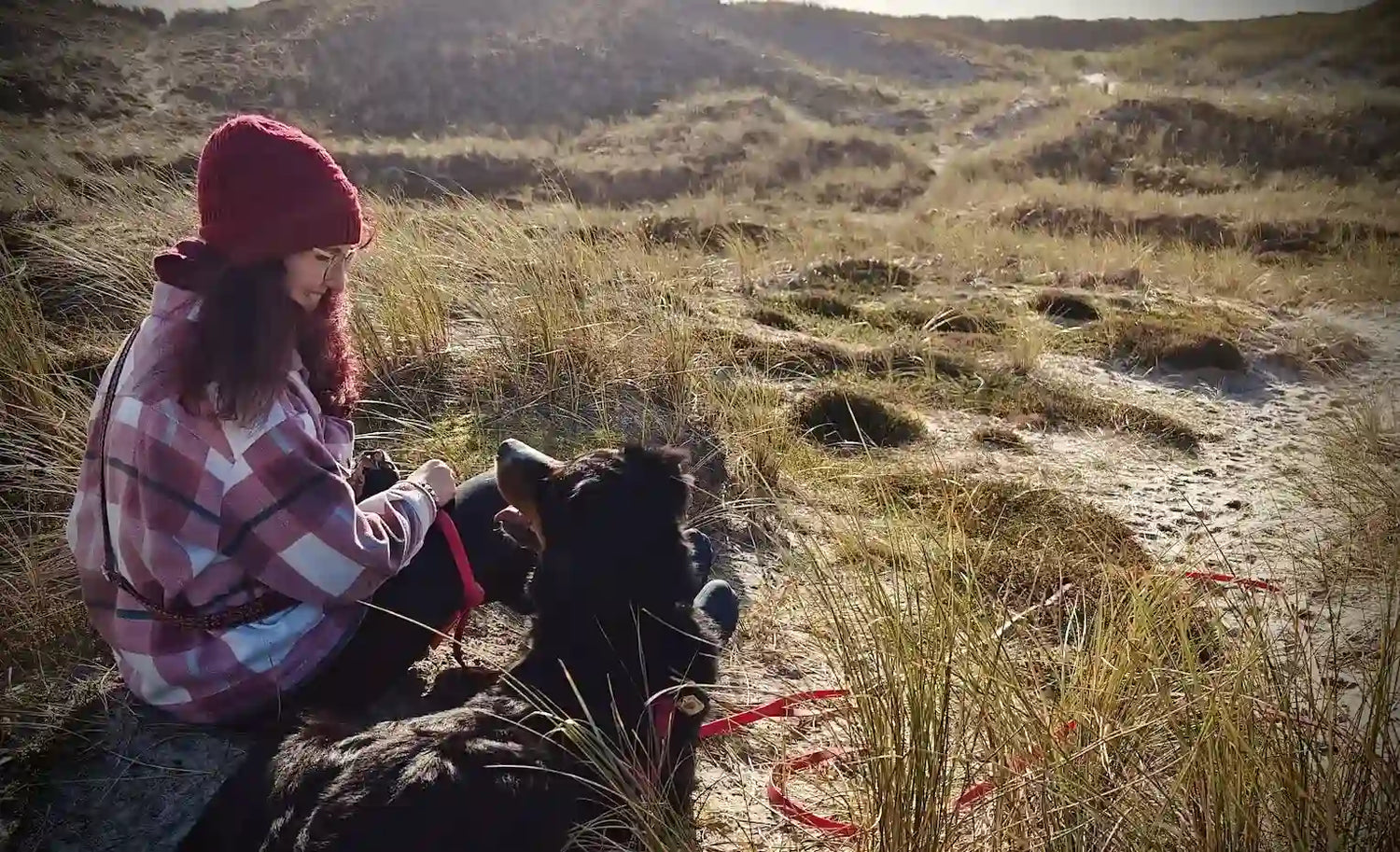 Person mit roter Mütze und karierter Jacke sitzt in den Dünen und hält eine Leine, an der ein schwarzer Hund liegt.