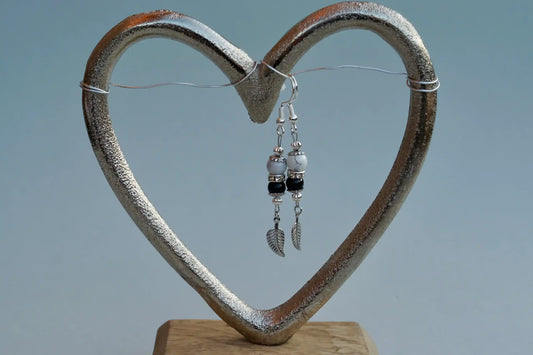 Ohrringe mit silberfarbenen Haken, zwei Edelsteinperlen in Magnesit und Turmalin sowie einem silberfarbenen Blattanhänger aufgehängt an einem herzförmigen Metallständer.