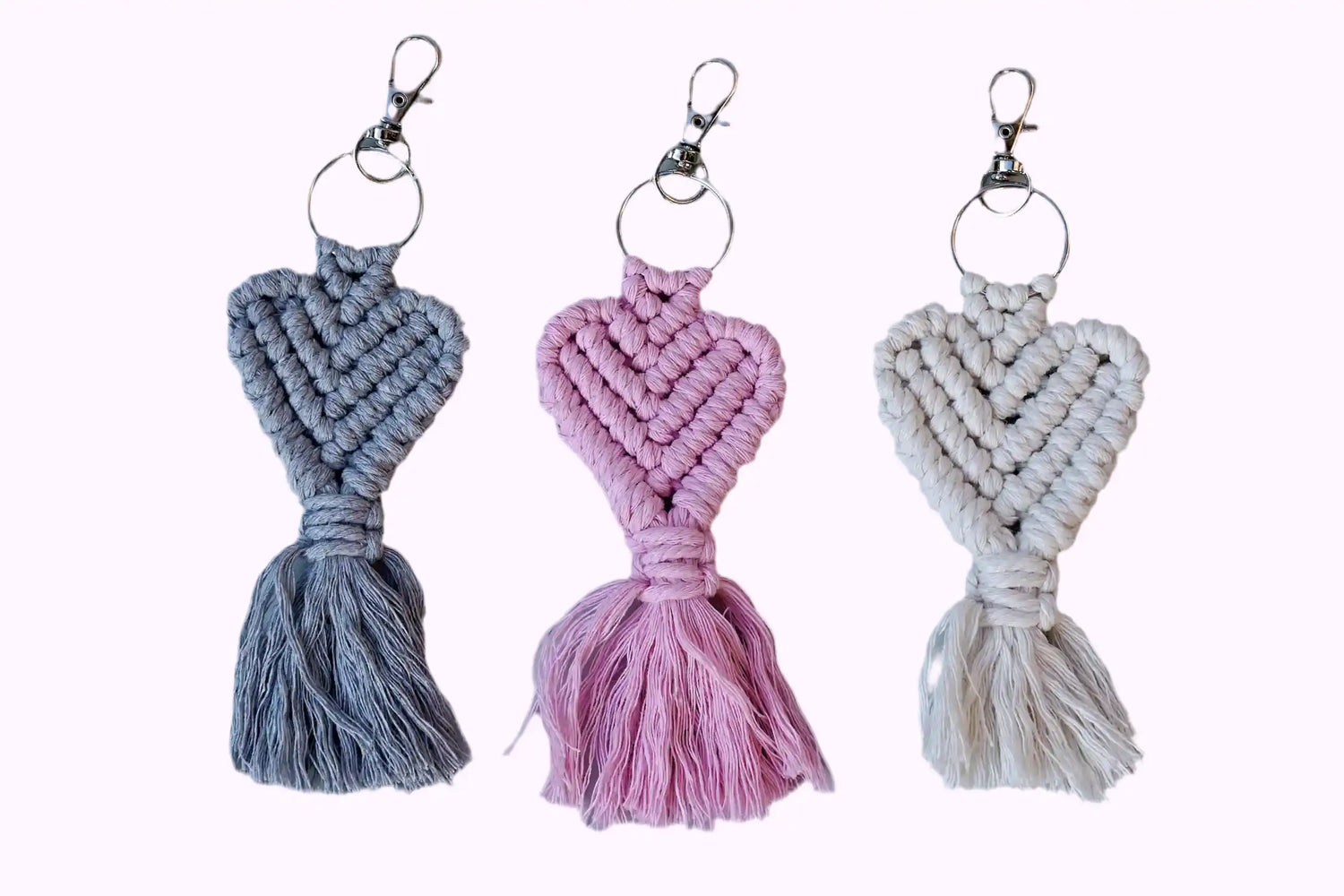 Drei Makramee-Schlüsselanhänger in Grau, Rosa und weiß mit Herzknotenmuster und Fransen, jeweils mit Metallring und Karabinerhaken