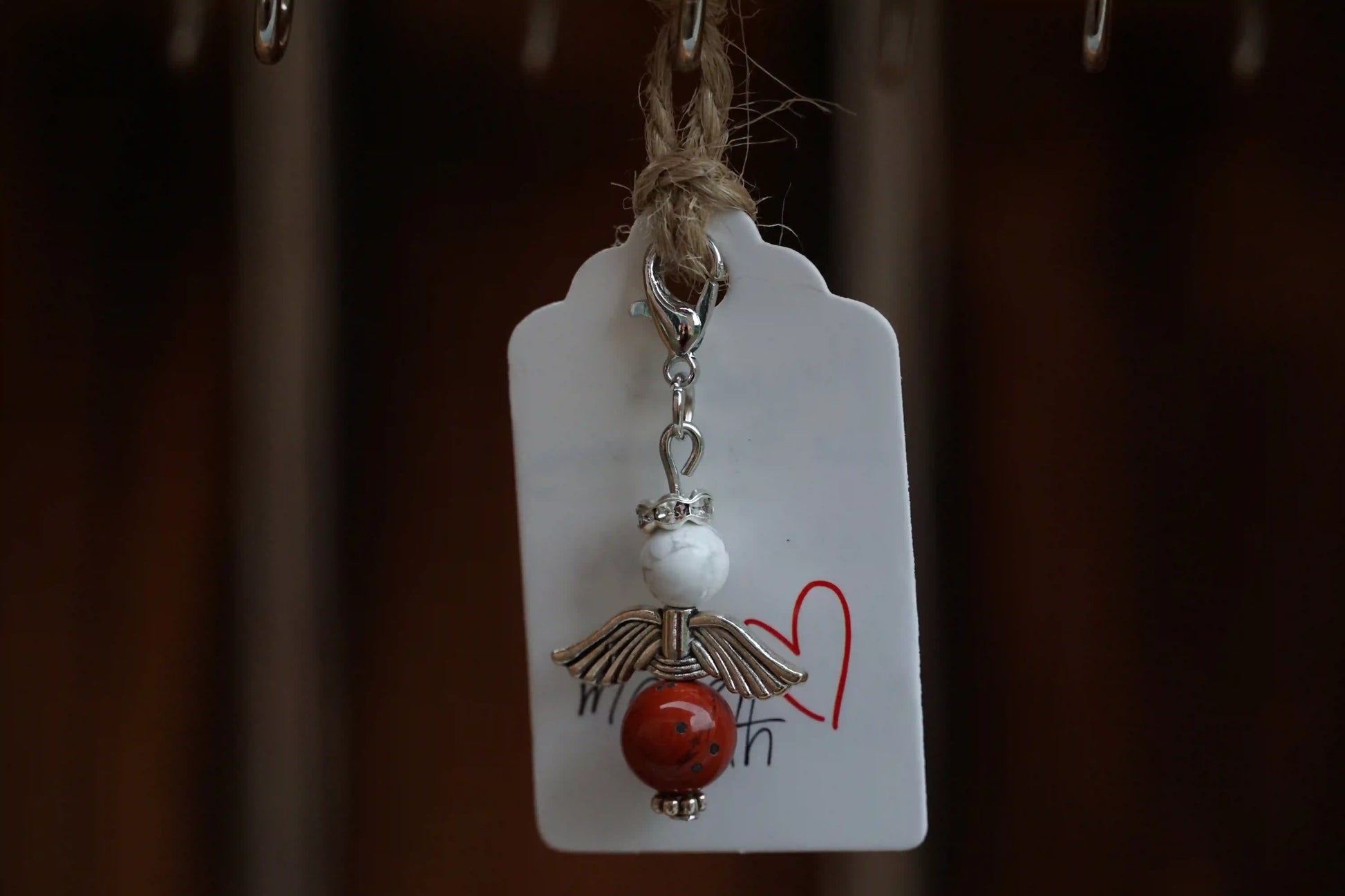Anhänger in Form eines Schutzengels mit Magnesit- und roter Jaspisperlen, silberfarbene Flügel, an einem Band befestigt.